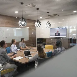 Fem mennesker sitter rundt en bord i et møterom og følger med på en skjerm.